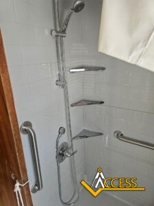 Accessible shower with grab bars, handheld and overhead showerheads, and three corner shelves. A curtain is partially visible on the right, and the Access logo appears in the bottom right corner.