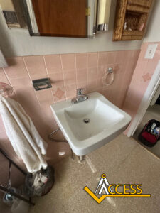 A small white wall-mounted sink with a towel hanging nearby, set against pink tiled bathroom walls. A trash bin and cleaning supplies are visible beneath the sink. The word ACCESS appears at the bottom right.