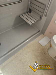 Accessible shower with a fold-down seat, grab bars on the wall, and a non-slip floor. A toilet and a logo that reads ACCESS are visible in the lower right corner.