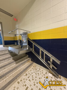 A wheelchair platform lift is installed on the side of tiled stairs with yellow, blue, and white walls. The lift is folded, with metal rails running alongside the staircase. The word ACCESS appears in the image corner.