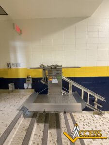 A platform wheelchair lift installed on a tiled staircase with yellow and blue stripes on the wall. The lift is unfolded, ready for use. The Access company logo is in the bottom right corner.