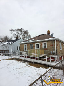 A small house with a stone exterior and a wheelchair ramp leading to the back door, surrounded by a snowy yard and a chain-link fence. The sky is overcast and neighboring houses are visible. Access company logo at the bottom right.