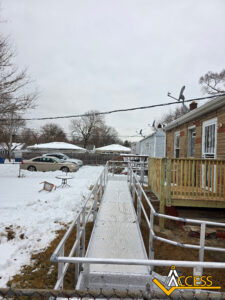 A metal wheelchair ramp leads from a wooden porch to a snow-covered backyard with scattered outdoor furniture. Houses and leafless trees are visible in the background under a cloudy sky.