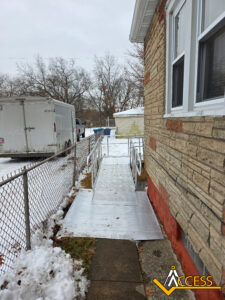 A metal wheelchair ramp leads from a sidewalk to the side door of a house. Snow covers the lawn and nearby trailer. A chain-link fence runs alongside the ramp. The ACCESS logo is in the bottom right corner.