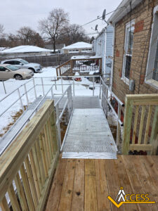 A metal wheelchair ramp with railings connects a wooden porch to a snowy backyard, providing accessible entry to a brick house. ACCESS company logo is visible in the lower right corner.