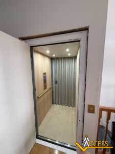 A modern residential elevator with wood-paneled walls and a metal accordion gate is open on an upper floor. The Access company logo is visible in the bottom right corner.