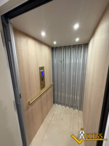 A modern residential elevator with light wood walls, a gold handrail, a control panel, overhead lights, and a collapsible metal gate. The Access company logo is visible in the bottom right corner.