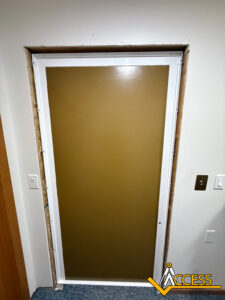 A brown door with a white frame is installed in a wall with exposed edges. Light switches and electrical outlets are visible on the wall. A logo reading ACCESS is in the bottom right corner.
