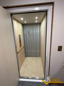A home elevator with wooden interior panels, a metal handrail on the left, a control panel, ceiling lights, and accordion-style metal gate, with the Access logo in the bottom right corner.