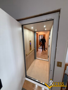 A residential elevator with light wood paneling and recessed ceiling lights is open on an upper floor. A man stands in the hallway outside. The Access Elevator logo appears in the bottom right corner.