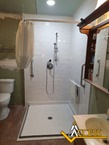 A white accessible shower with grab bars, a handheld showerhead, a fold-down seat, and a curtain. A toilet is on the left, and a wooden vanity with a mirror and shelves is on the right. ACCESS logo in the foreground.