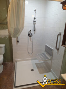 Accessible shower with white tile walls, a wall-mounted adjustable showerhead, a fold-down shower seat, a grab bar, a curtain, and a toilet nearby. The Access company logo appears in the lower right corner.