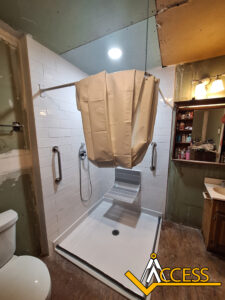 Accessible shower with white tile, a fold-up seat, grab bars, and a cream privacy curtain partially closed. The area appears under renovation, with exposed wall and ceiling sections. Logo “ACCESS” is in the bottom right.