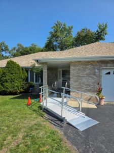 A brick house with a tan shingle roof has a silver wheelchair ramp leading to the front door. There are two orange cones near the ramp and green bushes and trees in the yard.