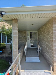 A concrete and brick entrance features a metal wheelchair ramp with railings leading to a white security door. A security camera, plants, and a window are visible on the left side.