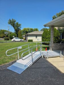 A metal wheelchair ramp with railings leads from a sidewalk to a building entrance, set next to a blacktop driveway. Two orange safety cones are placed beside the ramp, with grass, trees, and houses in the background.