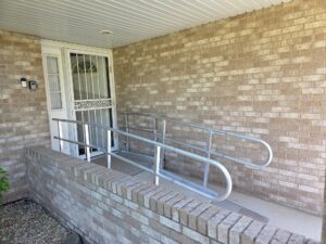 A metal wheelchair ramp with railings leads to a front door, surrounded by a beige brick wall and sheltered by a white overhang. A small garden area with gravel is visible on the left.