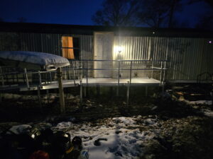 A metal wheelchair ramp leads up to the front door of a mobile home at night. The porch light is on, illuminating part of the ramp. Patches of snow and scattered items are visible on the ground.