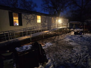 A mobile home at night with lights on inside and a metal wheelchair ramp leading to the entrance. Snow is scattered on the ground and objects are visible in the yard. Trees without leaves stand in the background.