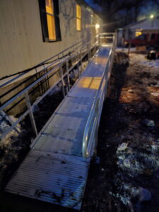A metal wheelchair ramp leads up to a mobile home at night, illuminated by light from a window. Snow and ice are scattered on the ground nearby.