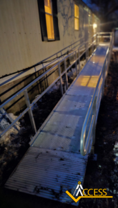 A metal wheelchair ramp with railings leads to the entrance of a building at dusk. Snow is visible on the ground, and warm light glows from the building’s windows. The word “ACCESS” is overlaid in the bottom right corner.
