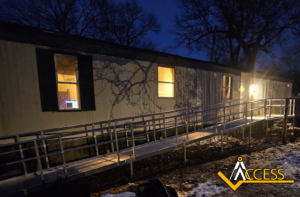 A modular building at night with illuminated windows and a lit accessibility ramp leading to the entrance. Bare trees cast shadows on the building, and theres snow on the ground. The ACCESS logo appears in the lower right corner.