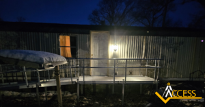 A mobile home with a lit porch light, visible ramp access, and a yellow Access logo in the corner. Snow is on the ground and the scene is set at dusk or early evening.