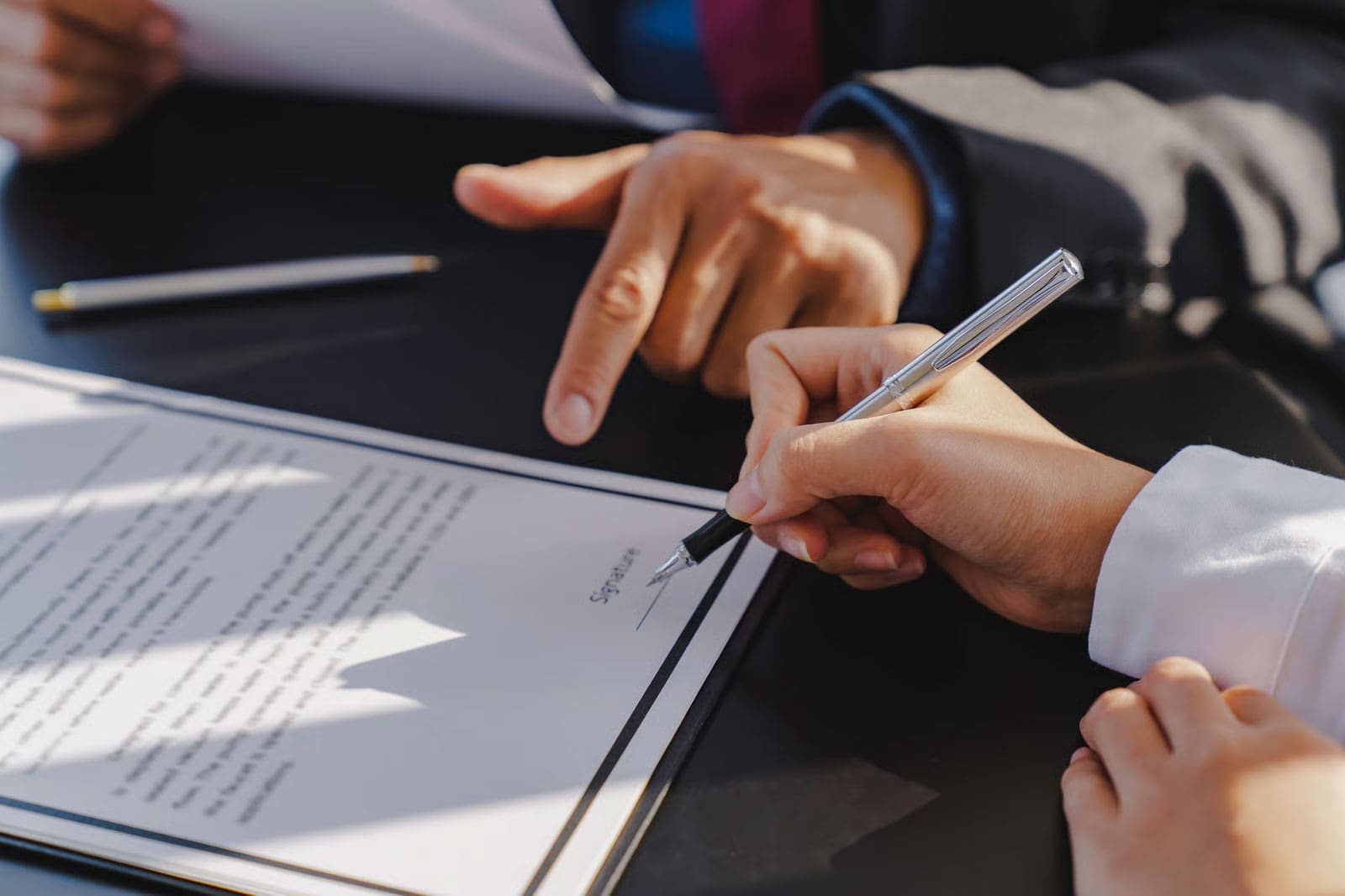 A person signs a document with a pen, while another in business attire points to the paper, creating an atmosphere of trust. A pen and another document are visible on the dark table.