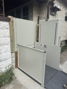 A residential vertical platform lift is installed outdoors by a house, next to a stone wall, providing accessibility for wheelchairs or mobility devices to bypass steps.