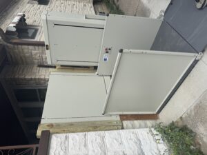 A residential wheelchair lift is installed outside a house, next to a stone wall and a pathway. The lift is in the lowered position, with control buttons visible on the side panel.