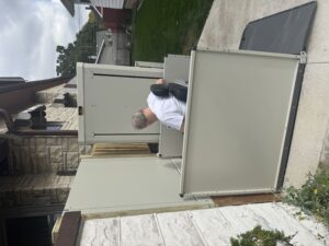 A person in a motorized wheelchair uses an outdoor vertical platform lift next to a house, providing accessible entry between the driveway and an elevated doorway.