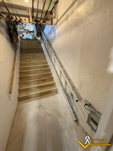 A wooden staircase in a basement with fishing rods mounted overhead and a long metal ladder stored along the wall. The Access logo appears at the bottom right corner of the image.