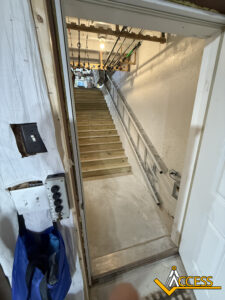 A doorway opens to a basement area with newly built wooden stairs leading up. A metal ladder is propped against the right wall, and pipes run along the ceiling. The Access logo appears in the bottom right corner.