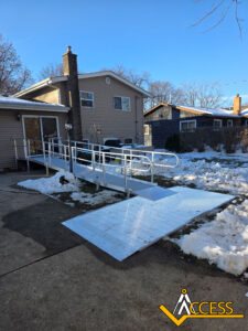 A silver metal wheelchair ramp leads from a snowy driveway to the entrance of a tan house. Snow covers the yard and parts of the ramp. The ACCESS logo is in the bottom right corner.