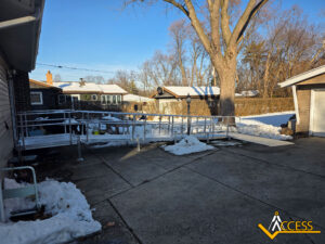A metal wheelchair ramp extends from a house to a driveway, surrounded by patches of snow. Trees and neighboring houses are visible in the background. The ACCESS company logo is in the lower right corner.