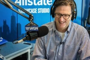 A man wearing glasses and headphones smiles while speaking into a microphone in a radio studio with WGN and Allstate Showcase Studio signs in the background, sharing tips on bathroom safety for independence at home.