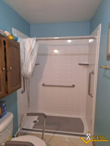 Accessible shower with white tiles, grab bars on three walls, a shower chair in front, a handheld showerhead hanging on the left, and a wooden cabinet partially visible on the left. ACCESS logo is in the bottom right corner.