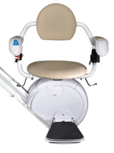 Front view of a Savaria K2 stairlift with the optional V seat, featuring a beige padded seat and open-back design, white armrests with integrated controls, a white carriage housing, and a black foldable footrest mounted on a straight stairlift rail. The V seat design provides additional seating space and improved comfort for some users.