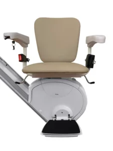 Front view of the Savaria K2 Plus stairlift with the L seat option, featuring a beige padded seat and backrest, adjustable armrests with integrated controls, a fold-down footrest, and a compact white carriage mounted on a straight stairlift rail. The traditional L seat design provides a comfortable seating position for residential or commercial stairlift applications.
