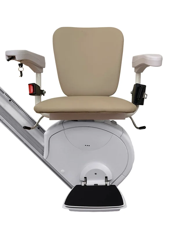 Front view of the Savaria K2 Plus stairlift with the L seat option, featuring a beige padded seat and backrest, adjustable armrests with integrated controls, a fold-down footrest, and a compact white carriage mounted on a straight stairlift rail. The traditional L seat design provides a comfortable seating position for residential or commercial stairlift applications.
