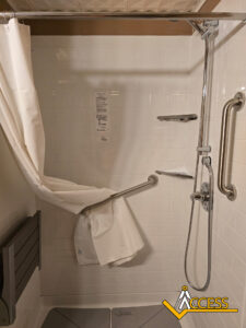 Accessible shower with white tile, a fold-down seat, grab bars, handheld showerhead, and a partially open curtain. The Access logo is visible in the lower right corner.