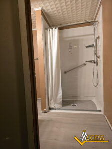 Accessible walk-in shower with a white curtain, grab bars, handheld showerhead, and a sloped floor for wheelchair access. The room has tan walls and a light-colored floor. The ACCESS logo is in the bottom right corner.