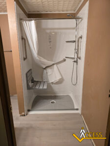 Accessible shower with grab bars, a fold-down seat, handheld showerhead, and a partially open curtain. The shower has a low threshold and non-slip floor, designed for easy access. The Acess logo is in the bottom right corner.