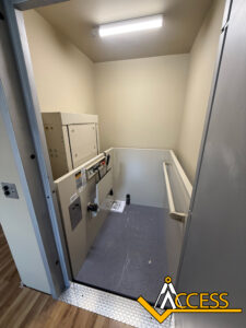 A small, indoor wheelchair lift with gray flooring and handrails on the right, located at the end of a hallway. The Access company logo is visible in the bottom right corner.
