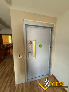 A gray elevator door with a small vertical window and stickers stands in a beige wall next to a wooden floor. A church interior with pews is visible through an open doorway on the left. The Access company logo is at the bottom right.