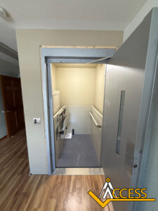 A small indoor wheelchair lift with a silver door open, revealing the platform and control panel inside. The space has light walls, wood flooring, and an Access logo in the bottom right corner.