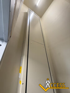 A tall, vertical beige elevator shaft inside a narrow room, viewed from below. Bright overhead light above. The Access company logo is at the bottom right corner.
