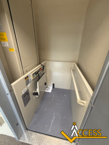 An empty wheelchair lift with safety rails on two sides, control panels, and instructions visible inside. The floor is gray, and there is a large Access logo in the bottom right corner.