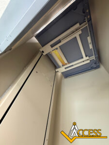 View looking up inside an open metal elevator shaft, showing part of the mechanical system above. The Access logo with a yellow check mark is in the bottom right corner.