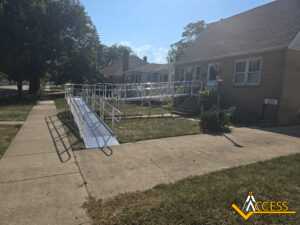 A wheelchair-accessible ramp made of metal extends from the sidewalk to the front entrance of a brick house. The ramp has railings on both sides and multiple turns across the yard. The sky is clear and sunny.